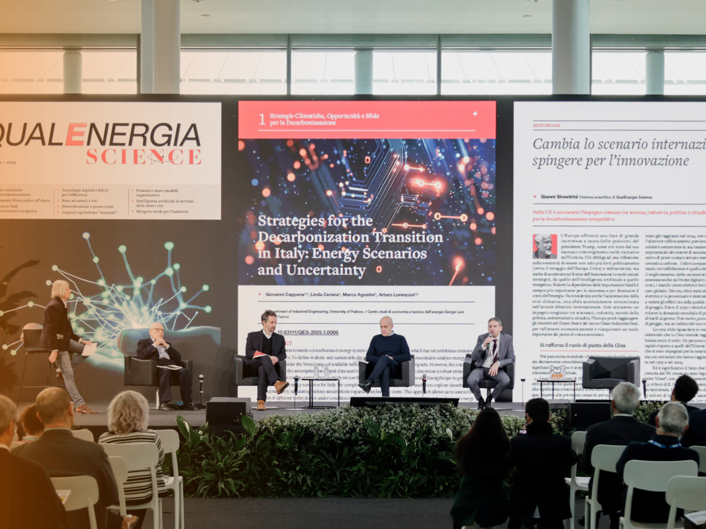 Panel di esperti sul palco di una conferenza sulle strategie per la transizione alla decarbonizzazione in Italia, sullo sfondo tre grandi schermi mostrano il logo della rivista 'QUALENERGIA SCIENCE' e un articolo intitolato 'Cambia lo scenario internazionale, spingere per l’innovazione'